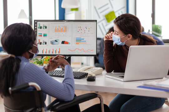 Freelancers wearing protective face masks working on computer in business office during global pandemic, disabled african woman sitting in wheelchair . Team discussing in new normal financial office.