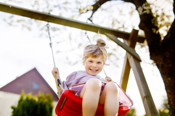 Happy little toddler girl having fun on swing in domestic garden. Smiling positive healthy child swinging on sunny day. Preschool girl laughing and crying. Active leisure and activity outdoors.