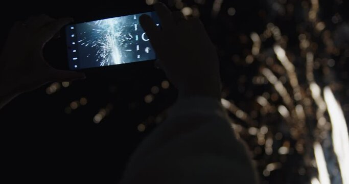 A woman capturing holiday fireworks at night with her phone during celebration.