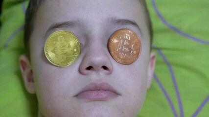 Teen Boy Lies, on a Bed with Gold Bitcoins on His Eyes. Humor. Child millionaire. Concept of mining cryptocurrency, wealth, blockchain, investment. 4K. Close up.