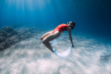 Sexy freediver woman in red swimsuit with white fins posing underwater over sand in ocean.