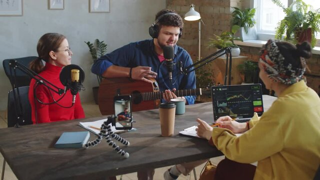 Young male musician in headphones playing the guitar and singing in mic, then smiling and giving interview to two female hosts in recording studio