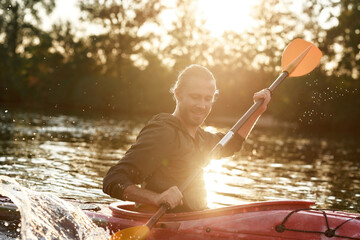 Caucasian young man looking cheerful, holding a paddle, kayaking in a lake surrounded by nature on a summer afternoon © Svitlana