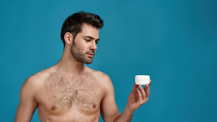 Fototapeta premium Portrait of naked model guy holding white jar with effective cream for face, posing isolated over blue background