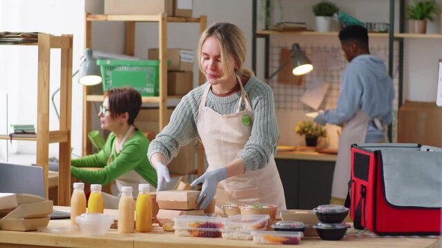 Young beautiful woman in apron and gloves taking notes on clipboard and packing paper containers with meals and bottled drinks in food delivery bag during workday in commercial kitchen