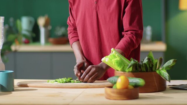 Tilt down shot of young Afro-American man cutting green salad leaves and putting them into blender while preparing healthy food in kitchen at home
