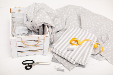process of sewing of bed linen. Sewing kit. Grey fabric, scissors, threads, measuring tape and white old wooden box on white wooden background