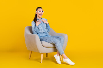 Full length body size photo woman sitting in chair amazed keeping hands on chest in glasses isolated vibrant yellow color background