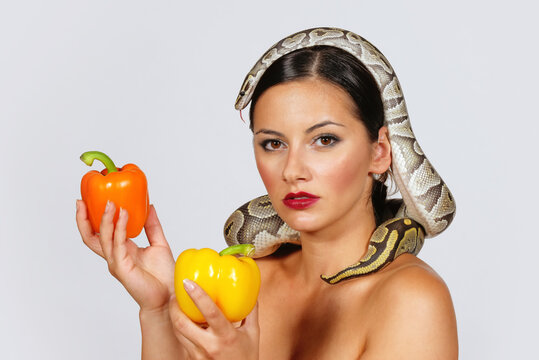 A beautiful young woman holds yellow and orange 
peppers in her hands while a python snake curls 
itself around her.