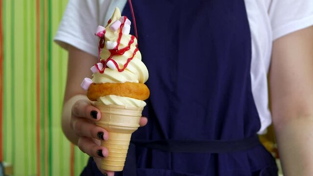 Strawberry topping is slowly poured onto ice cream in waffle cone. Salesman girl in blue apron holds delicious ice cream with marshmello and strawberry jam in her hands