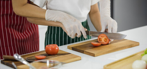 Unrecognizable woman teaching crop boy to cut ripe tomato on chopping board while preparing lunch in kitchen at home