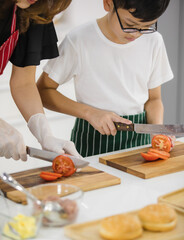 Unrecognizable woman teaching boy to cut ripe tomato on chopping board while preparing lunch in kitchen at home