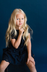 Naklejka premium beautiful blonde in a dark blue dress on a solid blue background. The photo is great for demonstrating the character of a child - daydreaming, enthusiasm, vulnerability, touching, beauty, stubbornness