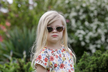 Portrait of fashionable toddler girl with long blonde hair in retro sunglasses and floral dress on a sunny summer day in park.	