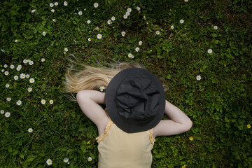 Outdoor portrait of toddler girl with long blonde hair laying down on a green meadow with hat over her face