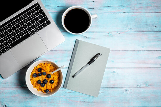 Open laptop computer with breakfast cereal with blueberries, notebook and pen and cup of black coffee for home working office or study on a weathered blue wood background