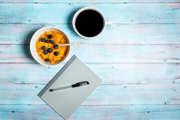 Notebook and pen with bowl of breakfast cereal with blueberries and cup of black coffee for home working office or study with copy space and room for text on a weathered blue wood background