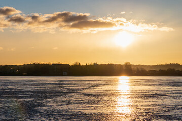Naklejka premium Winter landscape with frozen lake at sunrise or sunset. Lake glistening ice reflect a sun.Forest in the background.