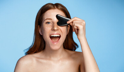 Smiling beautiful girl with closed eyes using facial gua sha jade board isolated on blue...