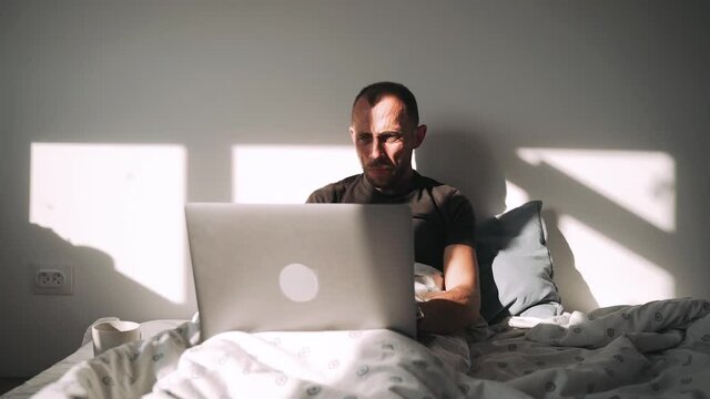 A man is lying on a bed and covered with a blanket working on a laptop A beautiful shadow from the sunset falls on a businessman who is working on a computer lying in bed Slow motion