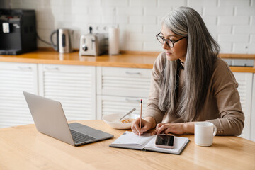 Mature grey woman writing down notes while working with laptop