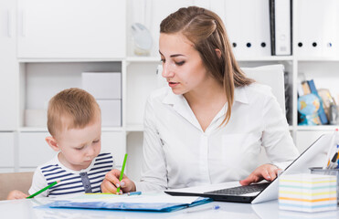 Woman is working on laptop while child painting on papers in office.