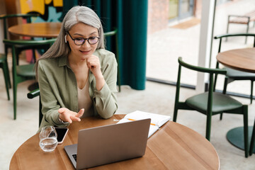 Mature smiling woman in earphone gesturing and using laptop