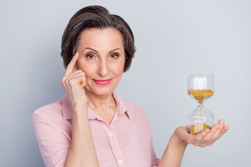 Photo of pretty thoughtful age woman dressed pink shirt holding sandglass finger head isolated grey color background