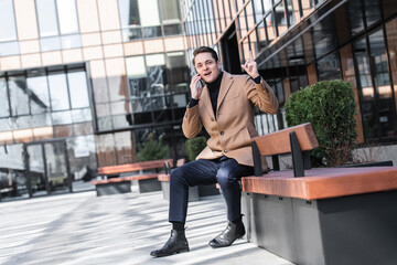 Young joyful man in business clothes sits on bench near modern business building, talking on phone.