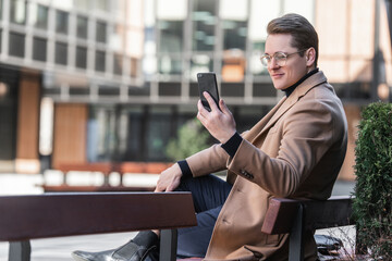 Young confident man with glasses sits on bench in courtyard of  business center, uses smartphone for video communication, video calls, online communication.