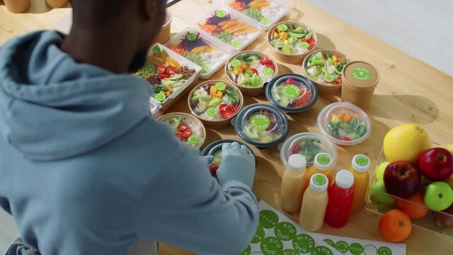 Over the shoulder high angle shot of black man in gloves putting stickers on containers with organic food while working at healthy meal delivery service