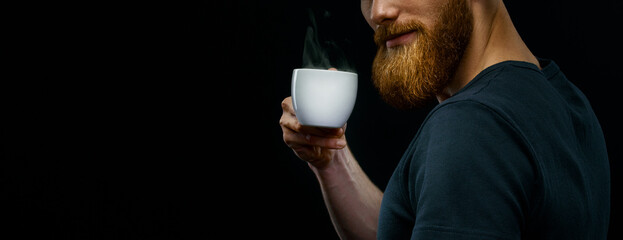 Cup of hot espresso coffee in the hand of a young bearded guy. Close-up studio shot on black background. Copy free space on left.