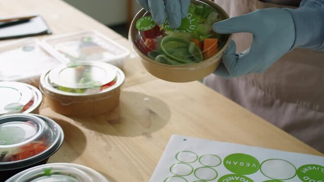Close up shot if unrecognizable worker in apron and gloves applying Gluten Free sticker on ecofriendly container with salad while working at healthy food delivery service