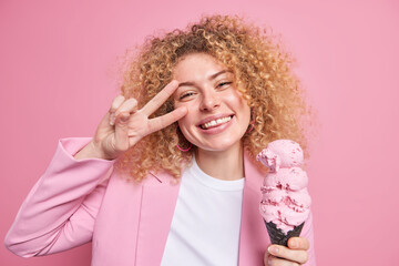 Positive pretty woman with curly hair eats huge ice cream in cone waffle makes peace gesture smiles broadly has fun during summer day dressed in stylish clothes isolated over pink background