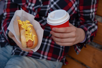 a hot dog and a red cup of coffee in the hands of an impersonal girl in a plaid shirt on a wooden...