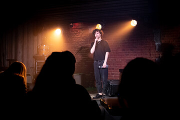 Young Caucasian male comedian performing his stand-up monologue on a stage of a small venue - obrazy, fototapety, plakaty