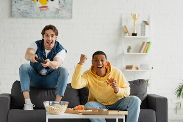 KYIV, UKRAINE - MARCH 22, 2021: interracial friends playing video game with joystick and enjoying pizza on couch in modern living room