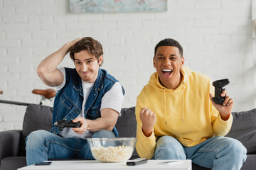 KYIV, UKRAINE - MARCH 22, 2021: interracial friends emotionally playing video game with joysticks in arms in living room