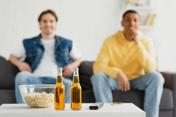 bottles of beer, TV remote and popcorn on blurred background with two interracial friends sitting on couch