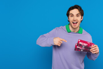 smiling young man showing concert tickets with point with finger gesture isolated on blue