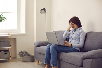 Tired unhappy woman covers face sitting on sofa at home. Employee stops working on laptop computer feeling exhausted and frustrated. Stress, burnout, life problems, chronic headache, eyestrain concept