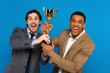 Happy interracial men in blazers holding trophy cup isolated on blue