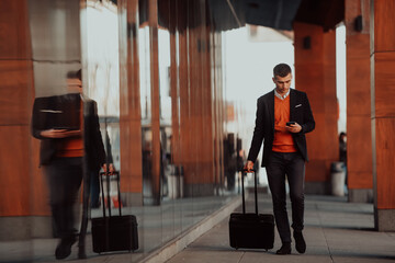 Fototapeta premium Going to airport terminal. Confident businessman traveler walking on city streets and pulling his suitcase drinking coffee and speaking on smartphone