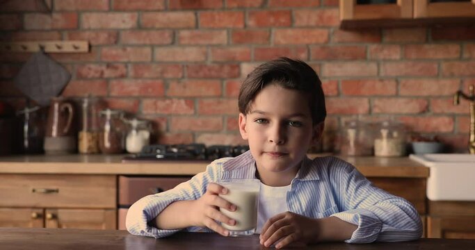 Little 7s boy sitting in kitchen looking at camera while drink glass of milk or sweet yoghurt protein vitamin source healthy beverage. Lifestyle healthcare, drink for health and growth of kid concept