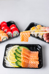Man hands holding raw salmon sashimi chopsticks. Lunch boxes, sushi set. Assorted rolls philadelphia, california, light background. Restaurant delivery, takeaway concept. Japanese cuisine seafood