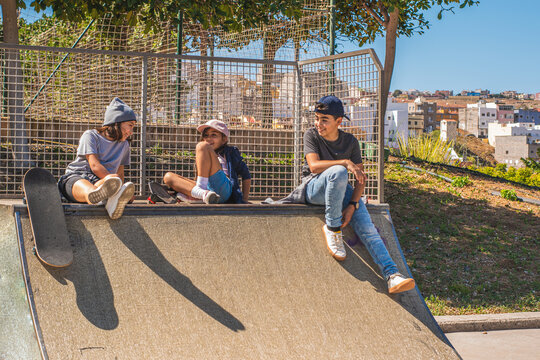 Three Child Skaters, With Their Skates, Sitting On An Obstacle On The Skate Park, Talking And Laughing Together.