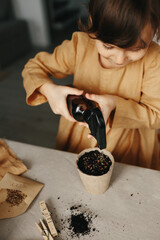 planting herbs at home. 6 years old girl wearing mustard linen dress watering plant in peat container with seed