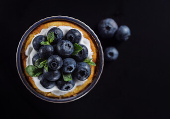 Blueberry tart in a ramekin with fresh berries and mint leaves. Homemade cheese cake at black background, top view.
