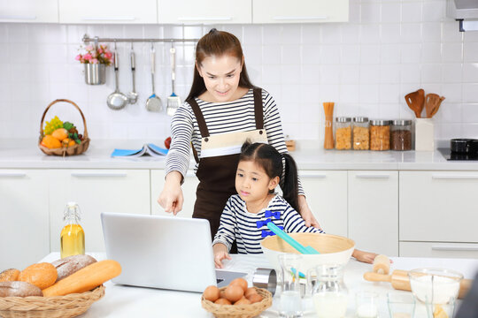 Happy Asian mother teaching her young daughter to bread baking in white modern kitchen by looking at recipe online from computer