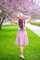 Beautiful woman in straw hat walking in a summer garden with blooming cherry trees. Girl wearing a pink long skirt